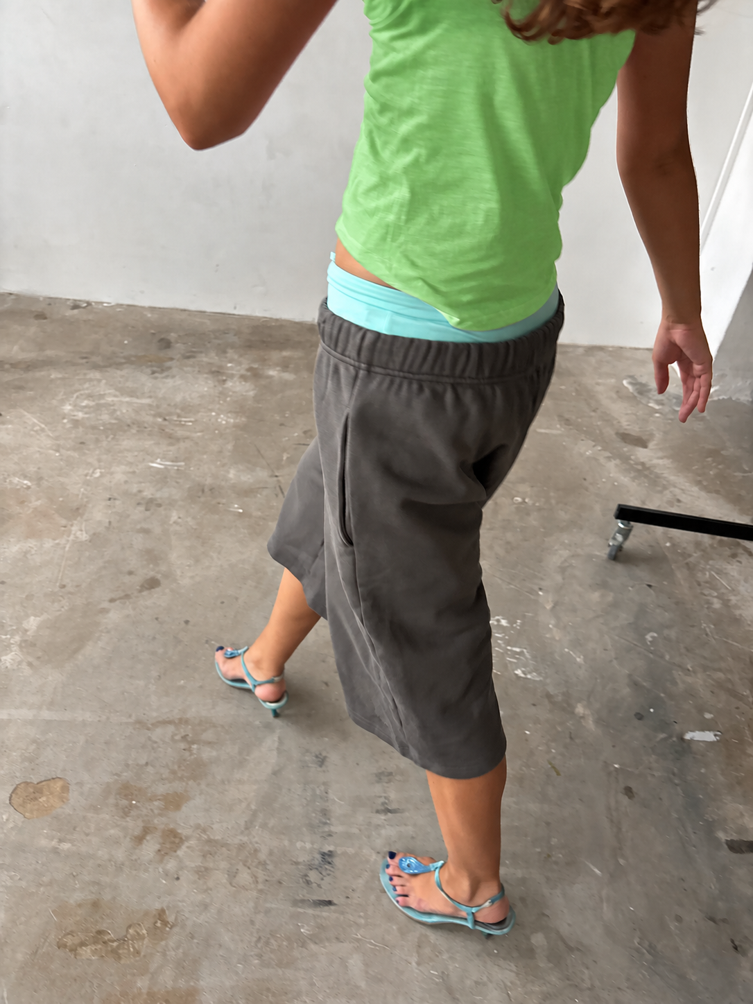 A person wearing dark gray knee-length shorts with a light blue undershirt peeking out at the waistband, paired with a lime green tank top and blue sandals, standing on a concrete floor.