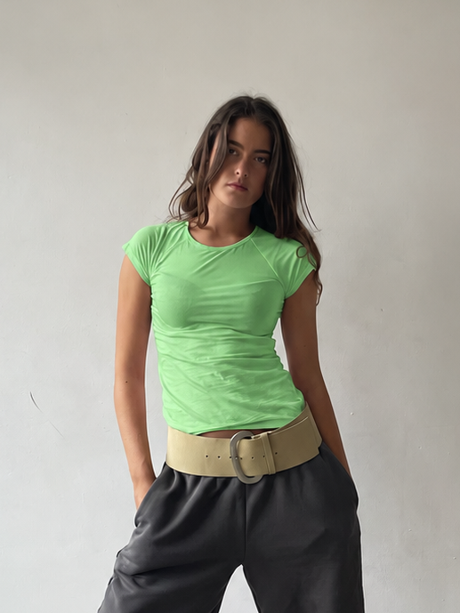 A woman modeling a lime green short-sleeve top, dark charcoal gray cropped pants, a beige belt, and white pointed-toe heels, standing against a plain off-white wall and concrete floor.