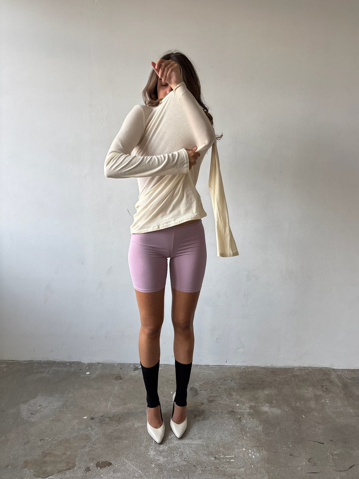 A woman stands in a minimalist room with a light gray wall and concrete floor, wearing a cream long-sleeve wrap-style top, lavender biker shorts, black knee-high socks, and white pointed-toe heels. She is holding the top open with one hand while pulling the fabric out into a long scarf-like drape with the other hand.