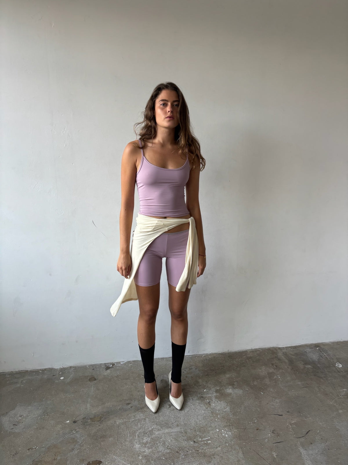 A woman stands against a plain light gray wall wearing a lavender ribbed tank top, matching lavender ribbed shorts, black knee-high socks, and white pointed heels, with a cream cardigan tied around her waist.