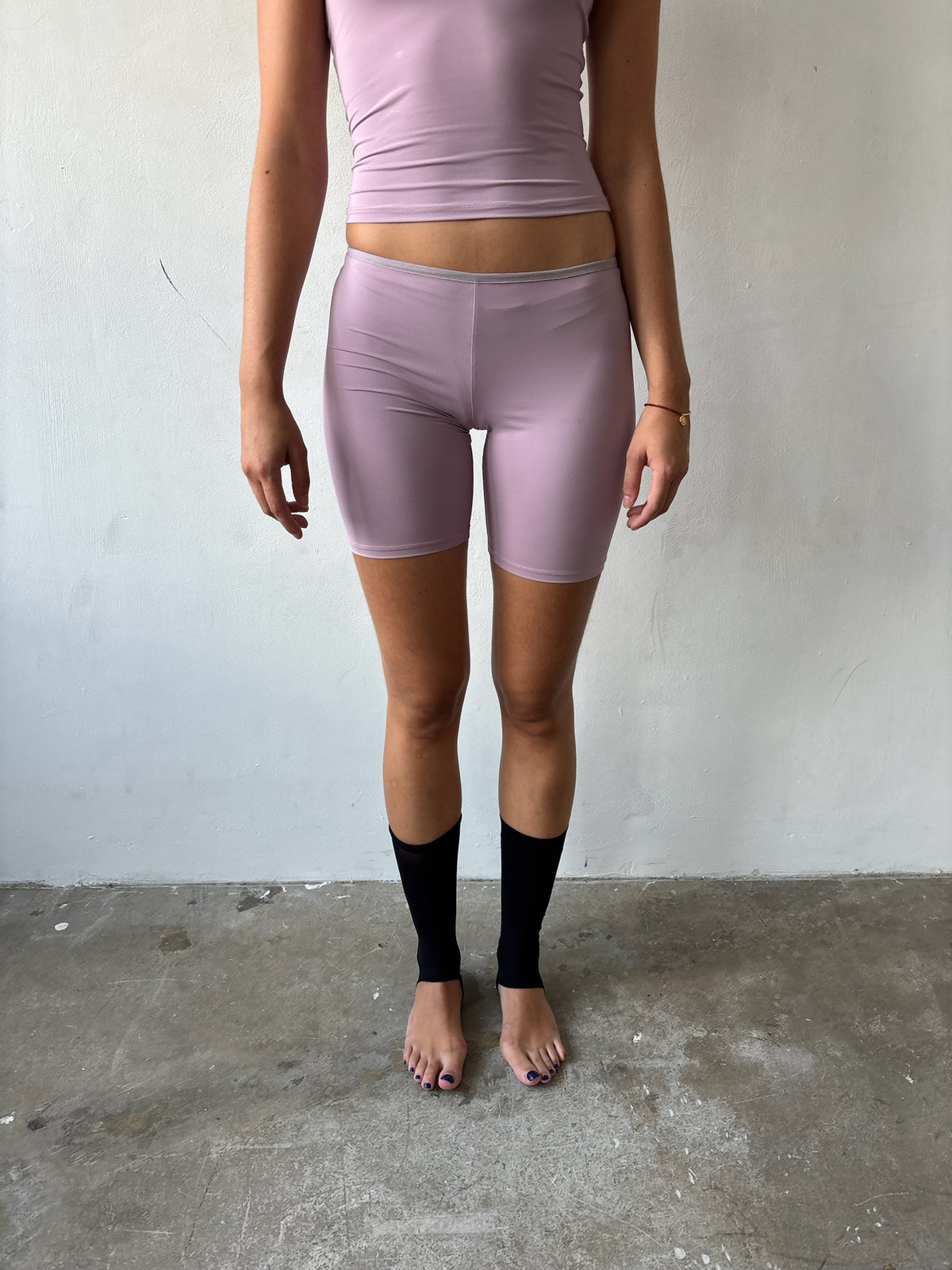 A model is shown from the chest down wearing light pink athletic biker shorts, a matching light pink crop top, and black compression socks, standing on a concrete floor against a plain off-white wall.