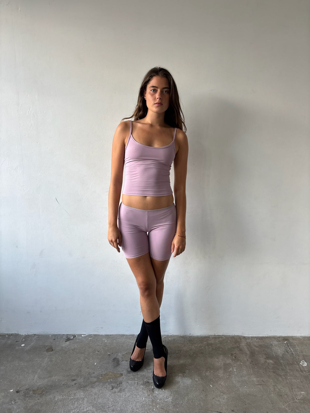 A woman stands against a plain off-white wall wearing a matching light pink/lavender ribbed camisole top and biker shorts set, paired with black socks and black heels.