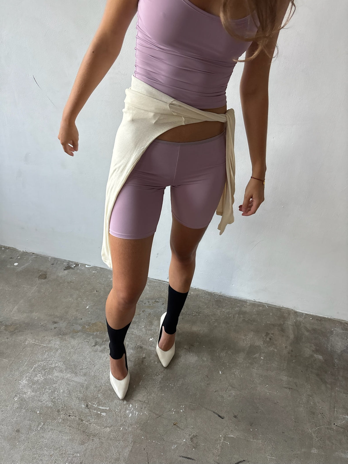 A model wearing a lavender ribbed tank top, matching lavender biker shorts, black knee-high socks, and white pointed-toe heels, with a cream knit wrap tied around her waist, standing on a concrete floor against a white wall.