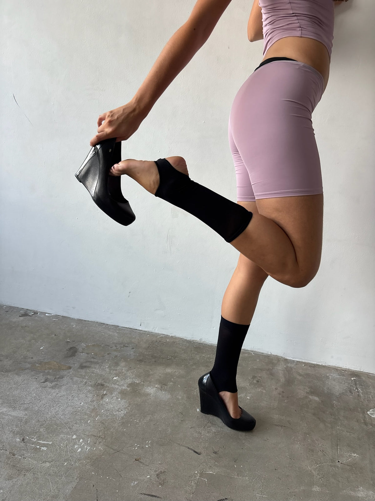 A model wearing a lavender athletic crop top and matching shorts, stretching their leg while wearing black knee-high socks and black Mary Jane wedge boots.