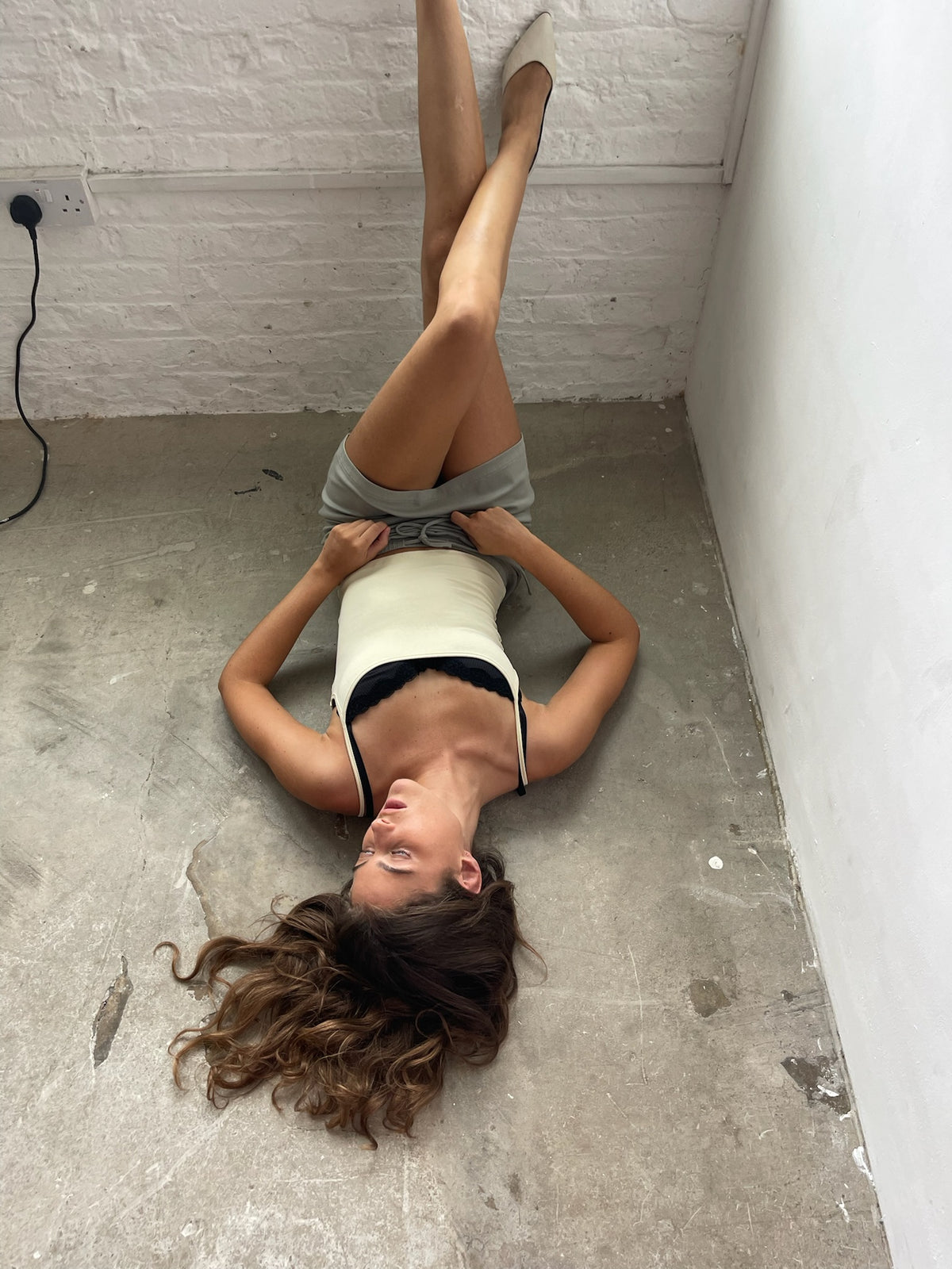 A woman lies on her back on a concrete floor against a white brick wall, wearing a cream and black bodysuit with gray shorts.