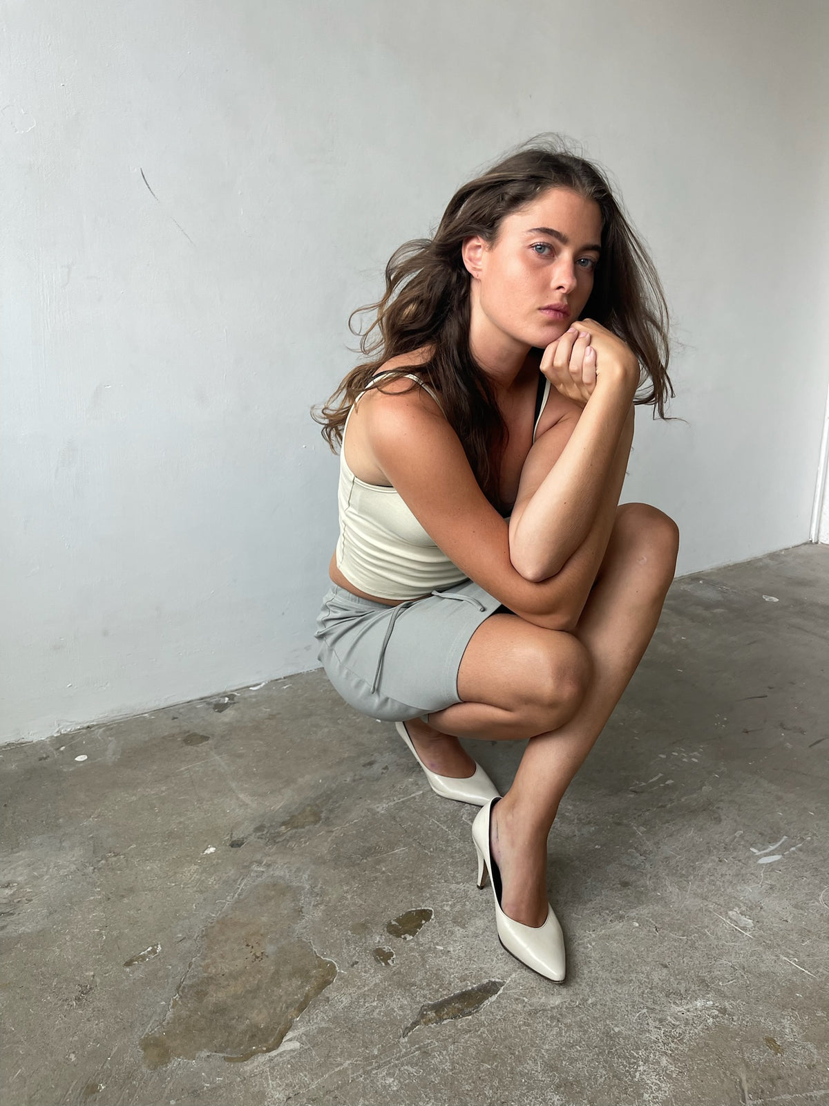 A woman crouching in a studio setting, wearing a light beige ribbed tank top, light gray mini skirt, and off-white pointed-toe heels.