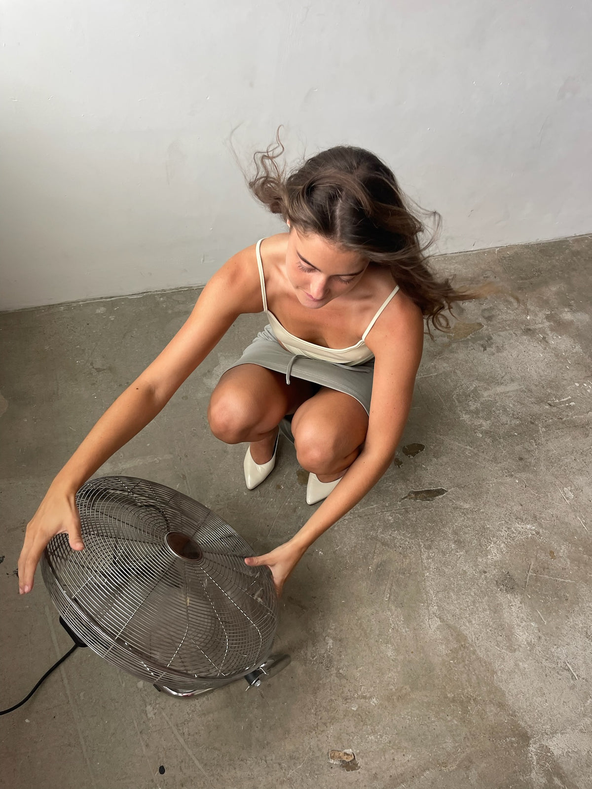 A woman crouches on a concrete floor against a plain wall, holding a large metal fan with a black cord.