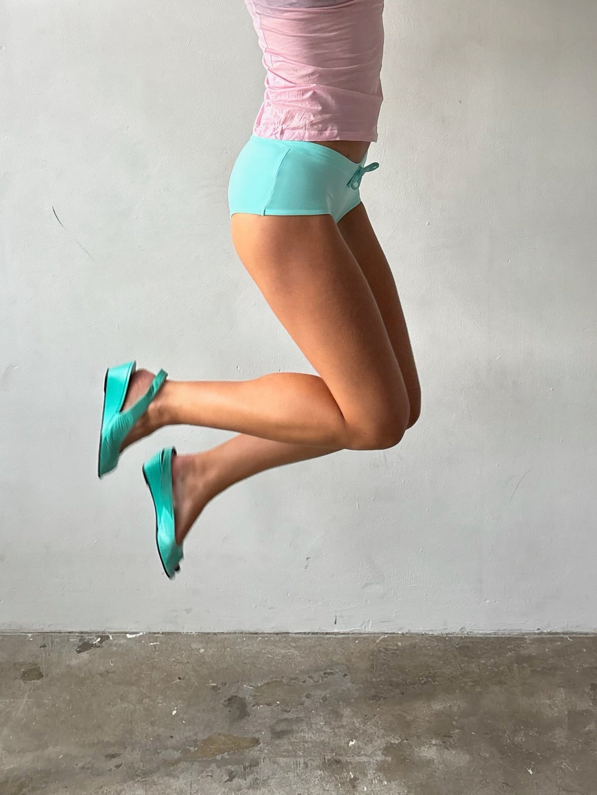 A person mid-jump wearing bright turquoise flat sandals with straps, light blue athletic shorts, and a pink top against a plain light gray wall and concrete floor.