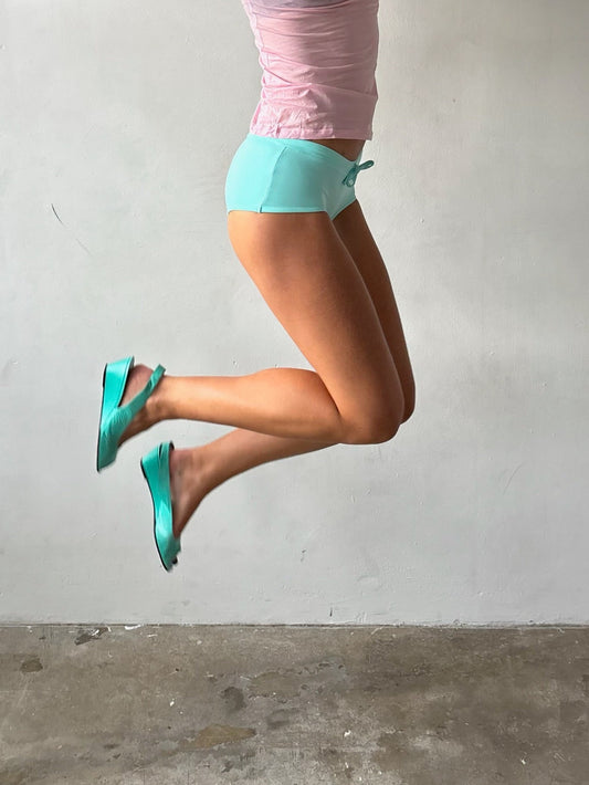 A person mid-jump wearing bright turquoise flat sandals with straps, light blue athletic shorts, and a pink top against a plain light gray wall and concrete floor.