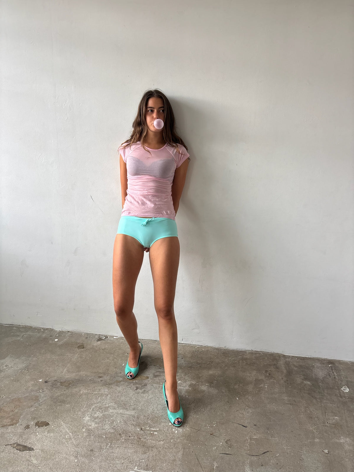 A woman stands against a plain off-white wall on a concrete floor, wearing a sheer light pink short-sleeve top, mint green high-waisted shorts, and mint green high heels.