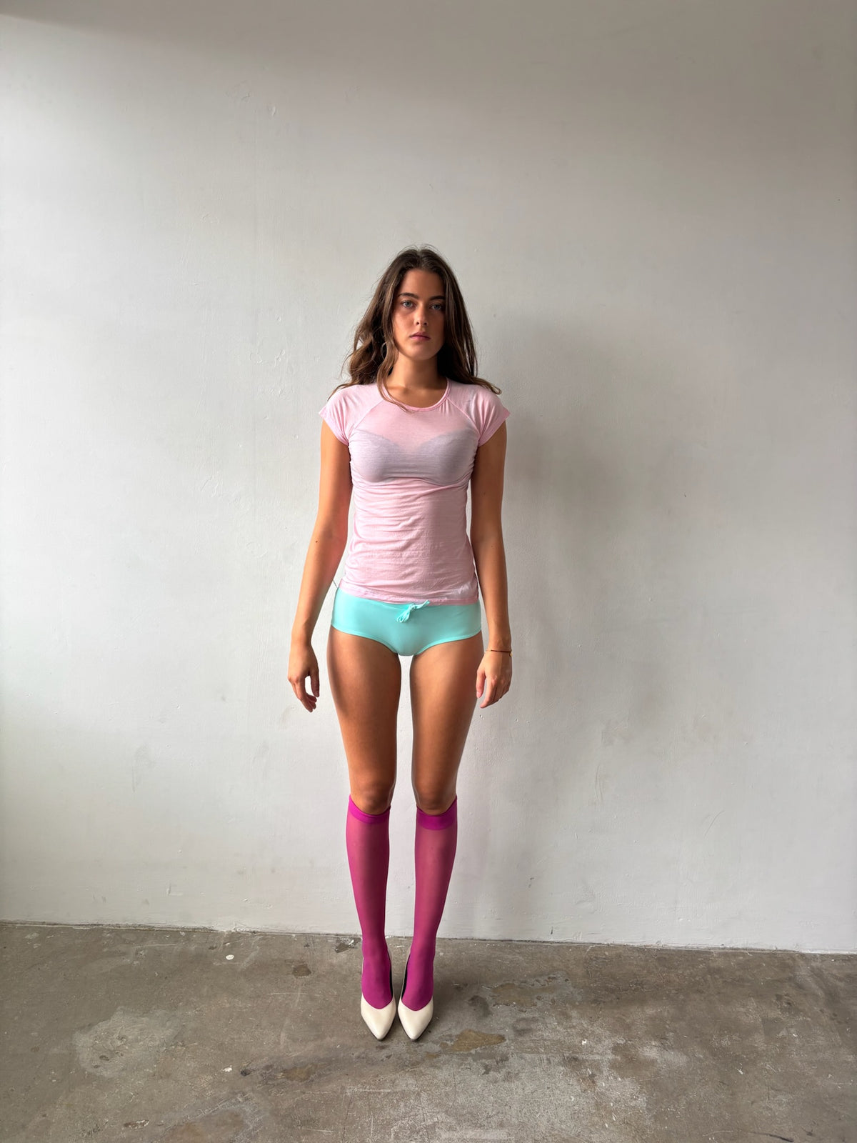 A woman stands against a plain off-white wall wearing a light pink short-sleeve top, mint green shorts, bright pink knee-high socks, and white pointed-toe heels.