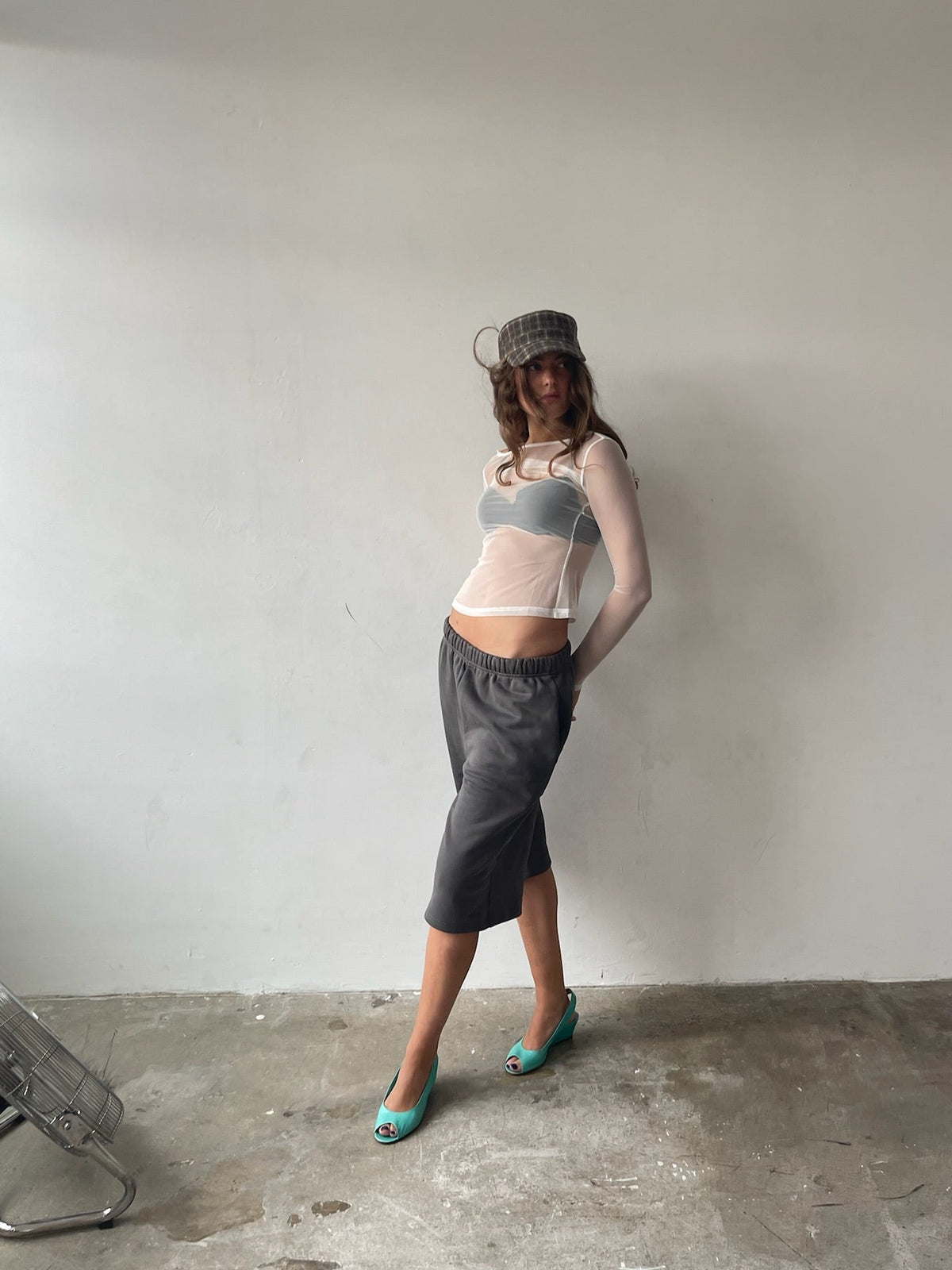 A woman stands in a minimalist room with a concrete floor and plain white wall, wearing a sheer white long-sleeve crop top, a gray knit skirt, a gray knit hat, and teal heels.