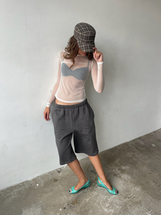 A woman stands against a plain light gray wall wearing a gray plaid flat cap, a sheer beige long-sleeve top over a gray bandeau, dark gray cropped pants, and mint green wedge flats.