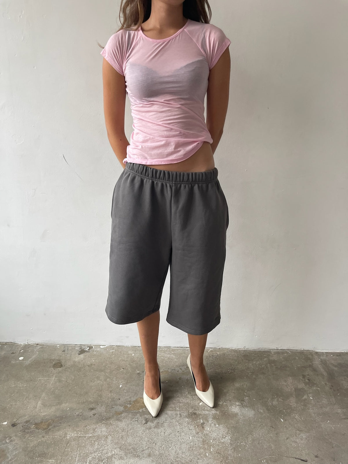 A model wearing a light pink short-sleeve t-shirt and dark gray cropped sweat shorts, paired with white pointed-toe heels, standing against a plain off-white wall and concrete floor.
