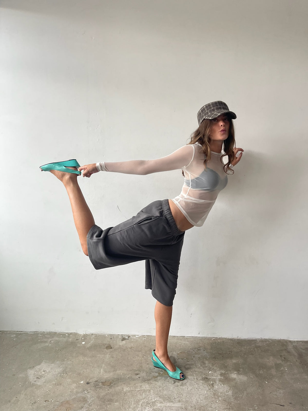 A woman in a grey cap, sheer long-sleeve top, dark grey wrap skirt, and mint green pointed flats poses in a high leg stretch against a plain white wall.