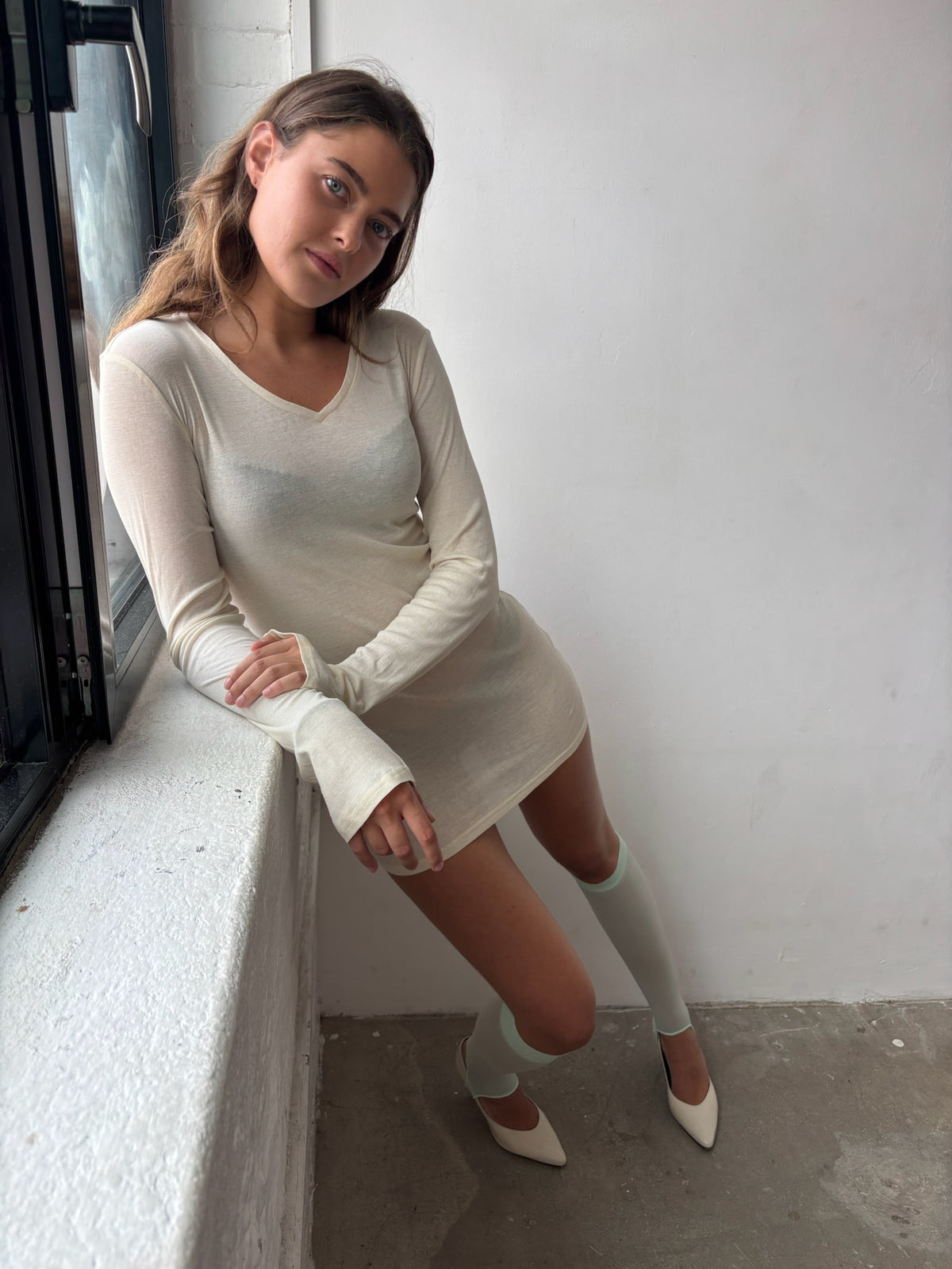 A woman wearing a cream long-sleeve mini dress, cream knee-high socks, and cream pointed-toe heels, leaning against a white wall next to a window.