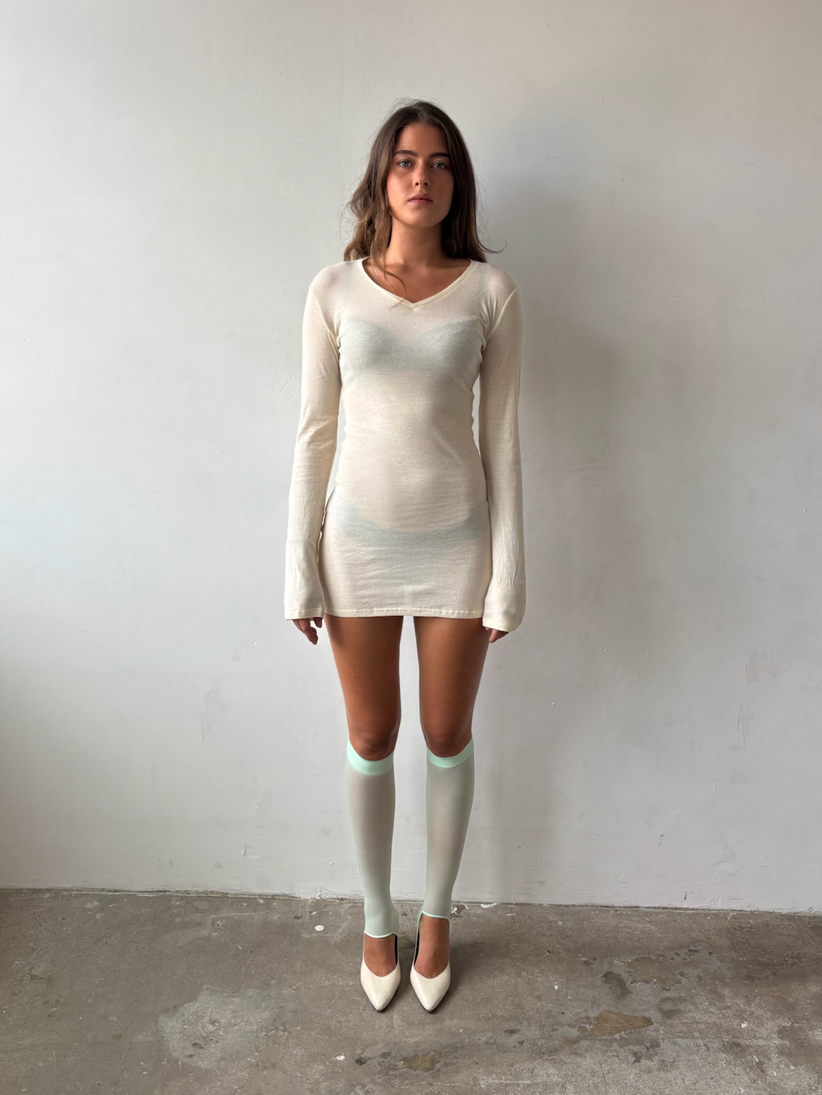 A woman stands against a plain light gray wall wearing a fitted cream long-sleeve mini dress with a sheer mesh overlay, light gray knee-high socks, and cream pointed-toe heels.