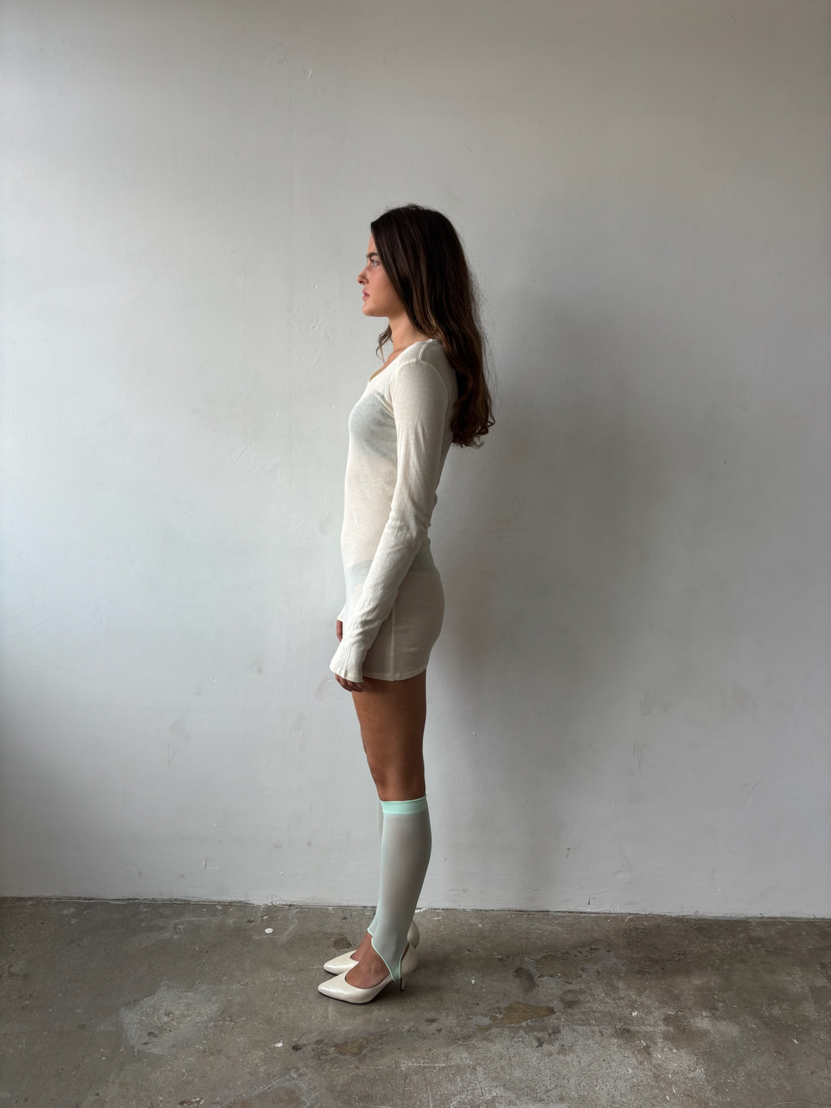 A woman stands in profile against a plain light gray wall and concrete floor, wearing a fitted off-white long-sleeve mini dress with a small front slit, light gray knee-high socks, and white pointed flats.