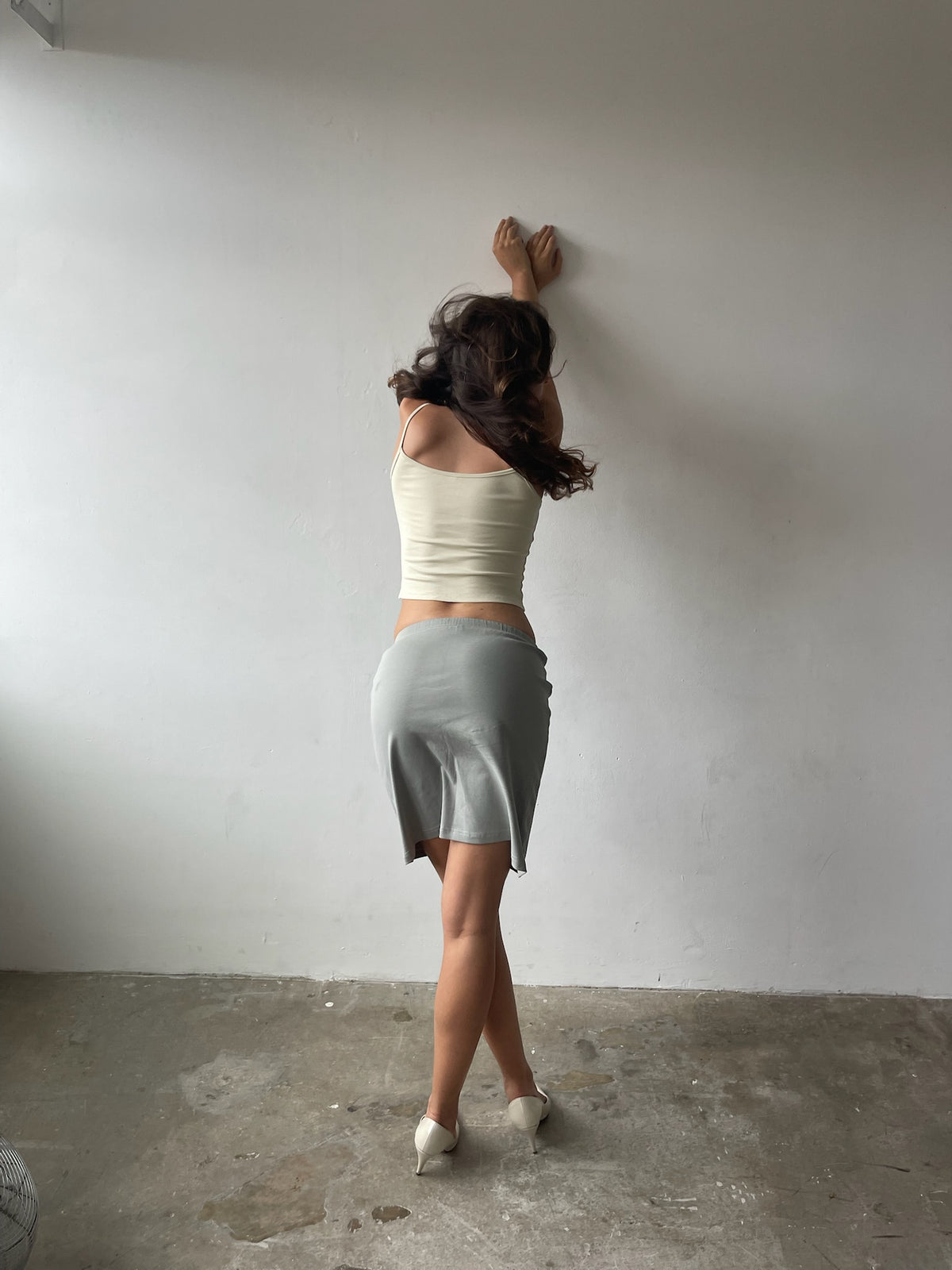 A woman stands with her back to the camera, leaning her hands against a plain white wall. She is wearing a cream off-the-shoulder crop top and a light gray pencil skirt, paired with white pointed-toe heels.