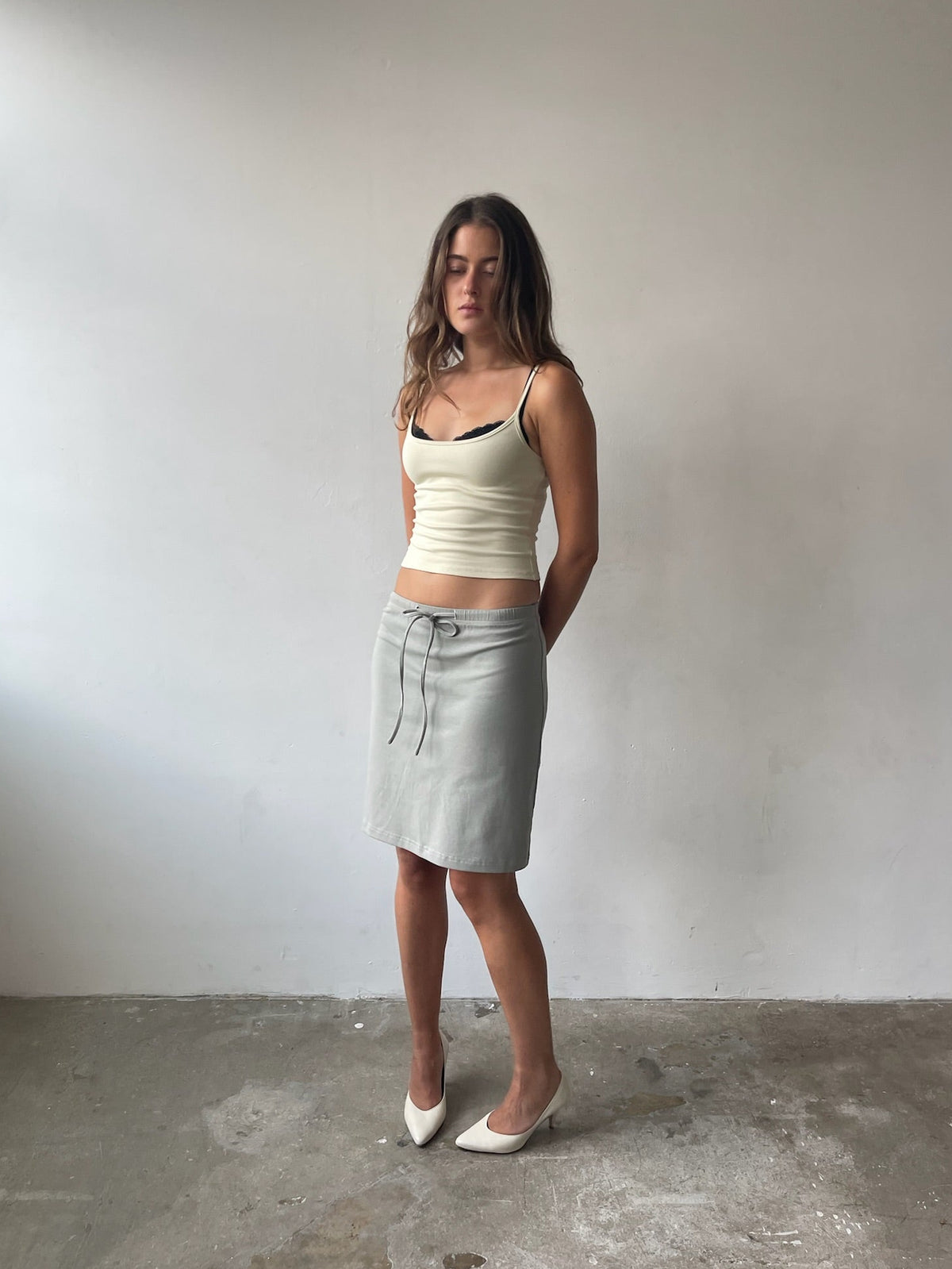 A woman stands in a minimalist room with a white wall and concrete floor, wearing a cream ribbed camisole with black trim and a light gray wrap-style pencil skirt with a drawstring.