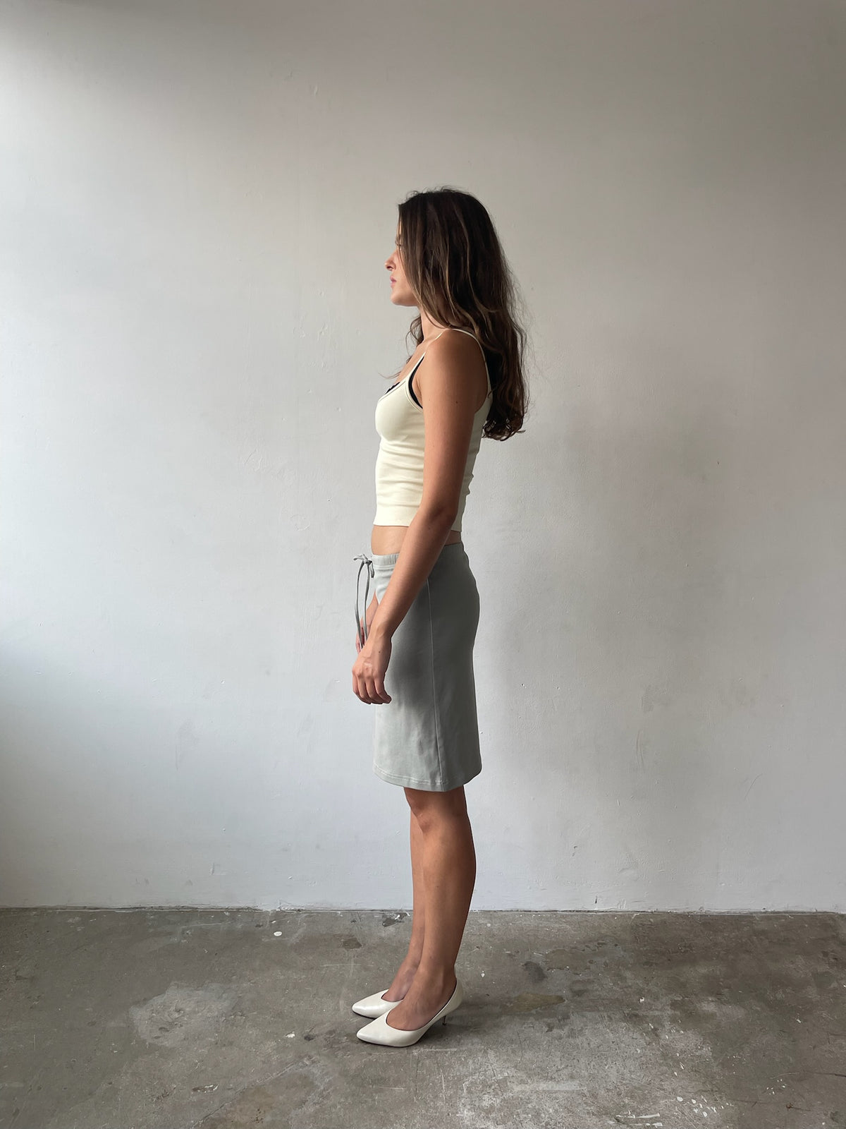 A woman stands in profile wearing a light beige ribbed tank top and a light gray knee-length skirt, paired with white pointed-toe flats, set against a plain gray wall and concrete floor.