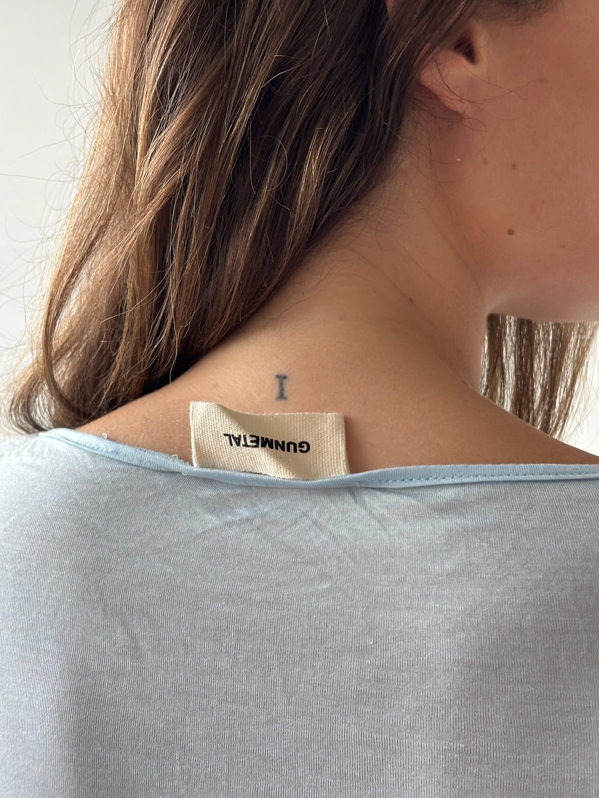 Close-up of a woman's neck and upper shoulder wearing a light blue top, showing a small black letter tattoo and a beige neck label that reads GUNMETAL.