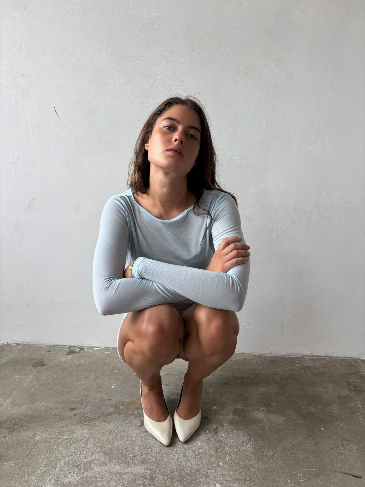 A woman crouching in a light blue long-sleeve top and white pointed-toe heels against a plain white wall and concrete floor.